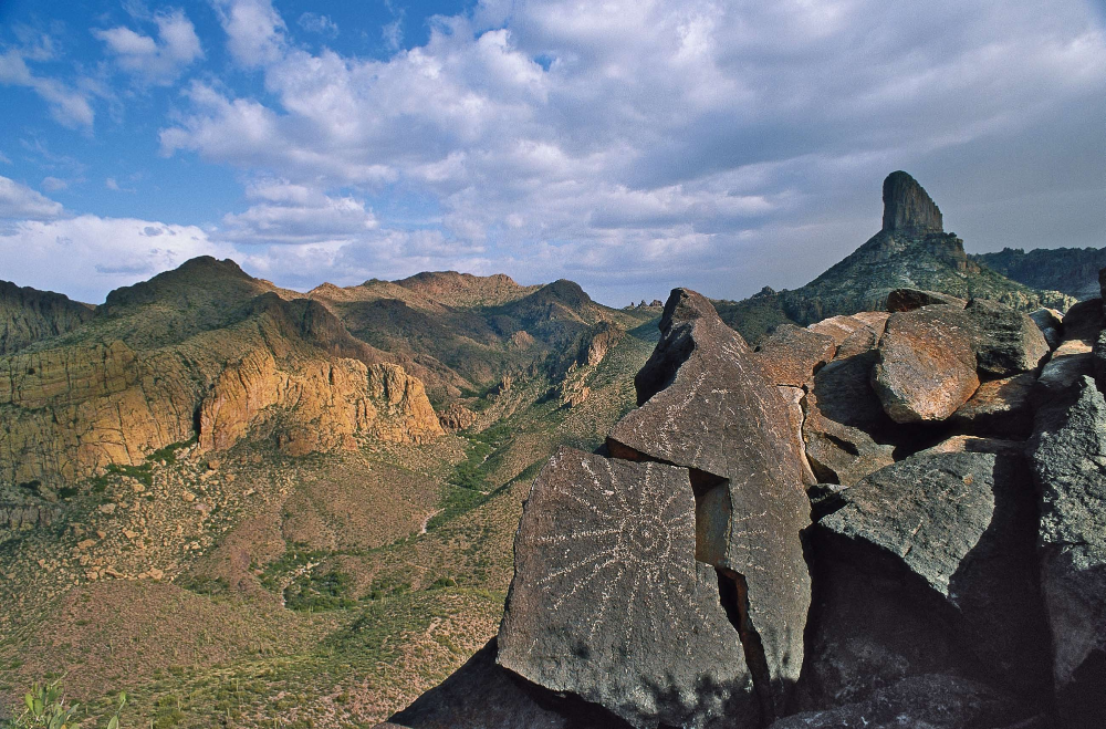 Etched in stones atop Black Top Mesa, a Spanish symbol points across the rugged terrain of the Superstition Wilderness to the distant Weavers Needle. Cast in shadow, the distinctive rock formation reigns over a long and dark history of gold, greed and murder.